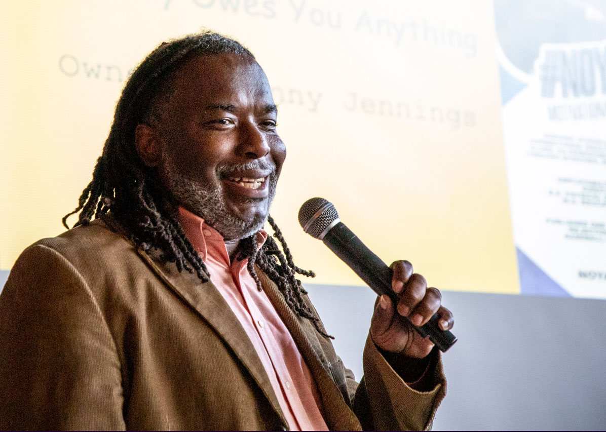 Man smiling, wearing a brown corduroy jacket, holding a microphone presenting his pitch at the Benton Harbor Pitch Competition